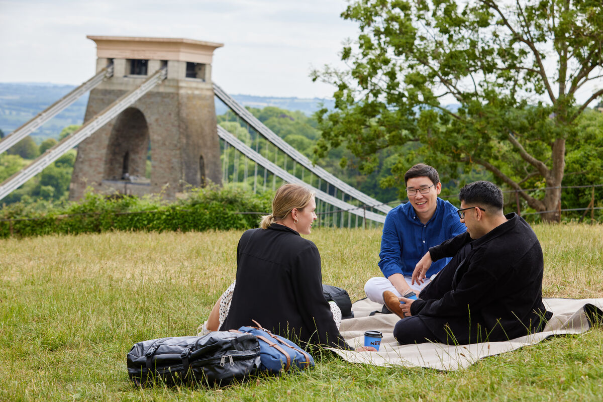 three students sitting on the grass with the suspension bridge in the background